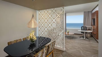 Una habitación de hotel moderna con comedor que incluye una mesa negra y sillas estampadas, un separador decorativo, una pequeña mesa redonda con una silla, un televisor montado en la pared y vista al mar.