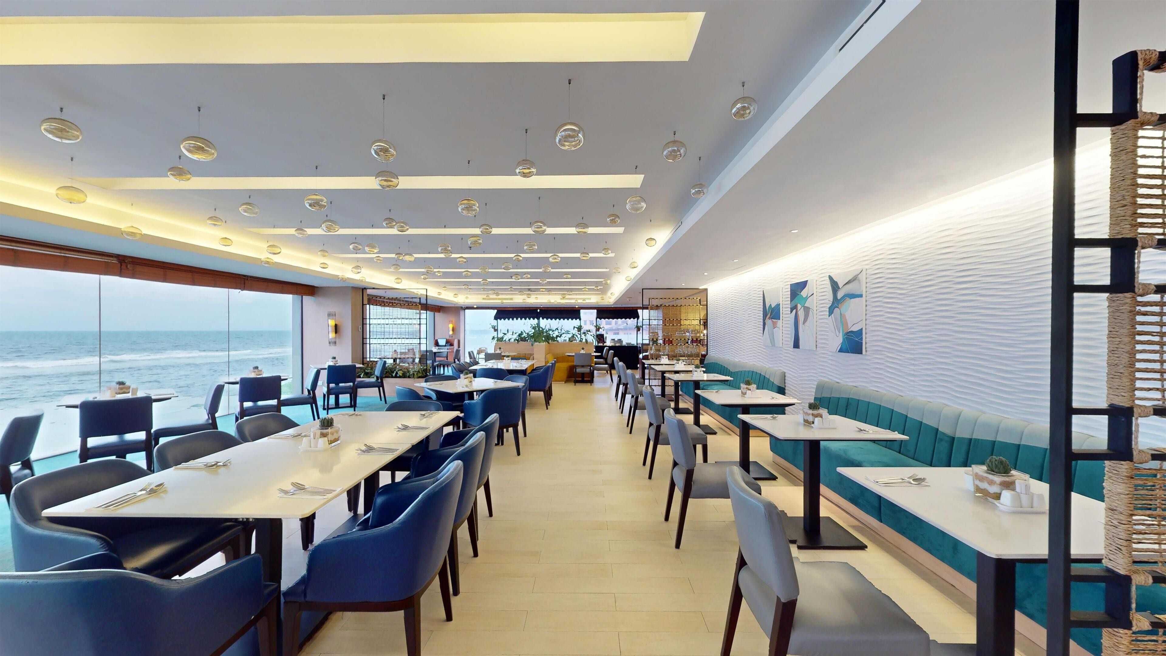 A restaurant with tables and chairs arranged along a wall with a view of the ocean.