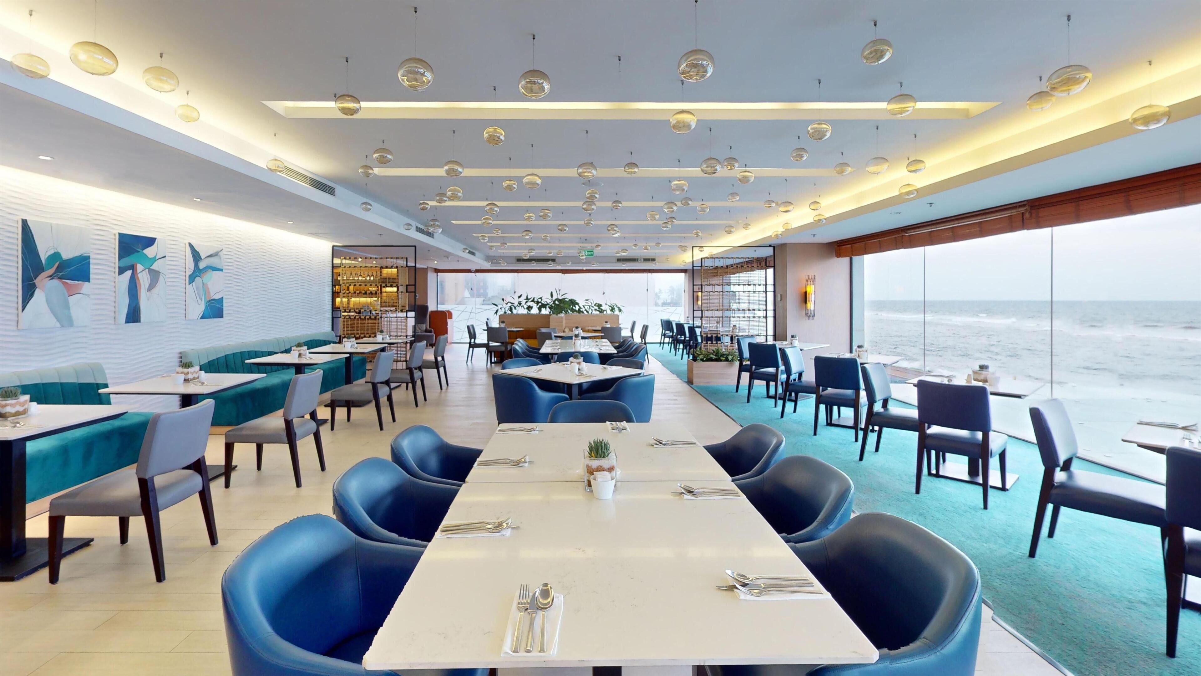 A modern restaurant with a long table in the center, blue chairs, and a view of the ocean.