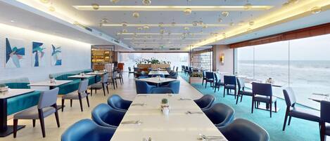 A modern restaurant with a long table in the center, blue chairs, and a view of the ocean.