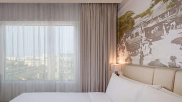 A hotel room with a large bed, a view of the city through sheer curtains, a wall with a textured mural, and a bedside lamp.