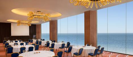 A conference room with round tables and blue chairs, large windows with a view of the sea, and ornate gold chandeliers.