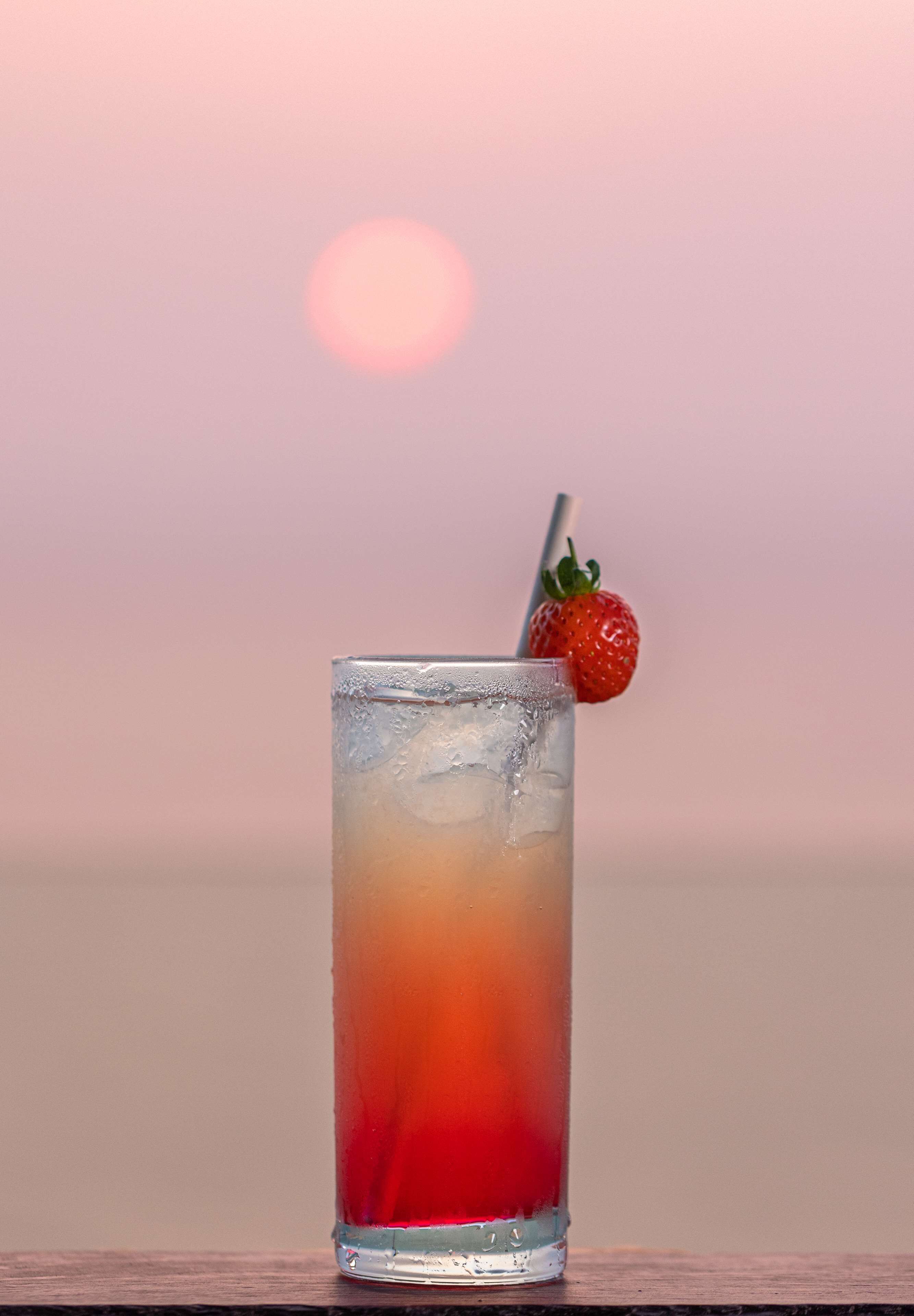 A tall glass with a gradient drink, ice cubes, and a strawberry garnish against a sunset background.