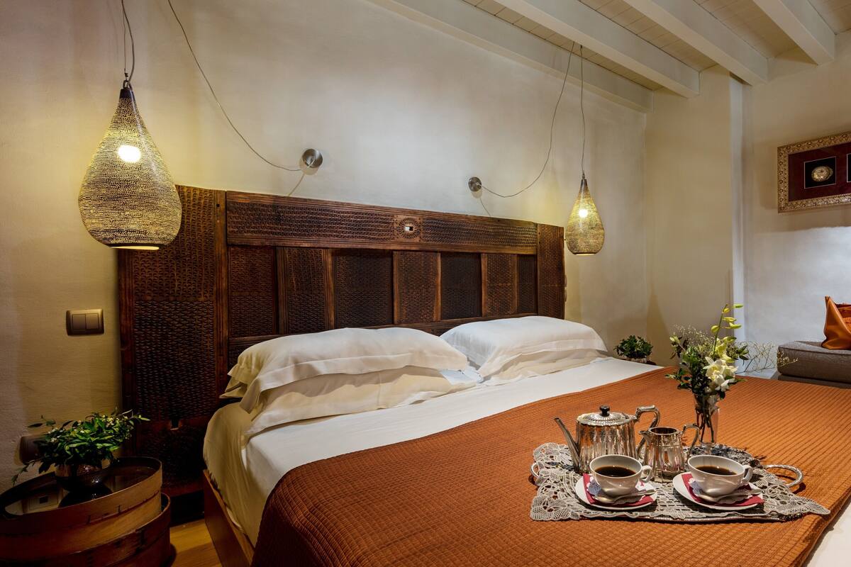 A neatly made bed with a brown quilt, a bedside table with a teapot and cups, and a wall-mounted decorative piece.