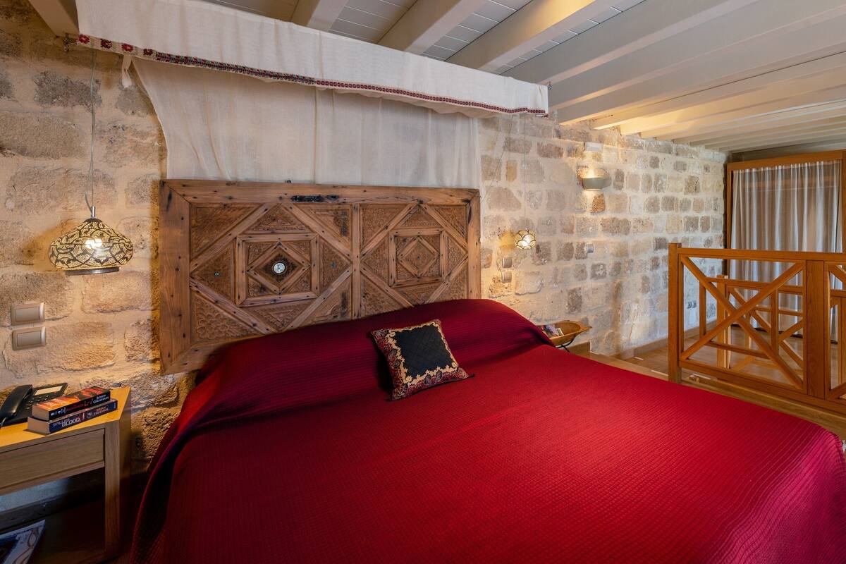 A bedroom with a stone wall, a wooden headboard, a red bedspread, and a wooden nightstand with books.