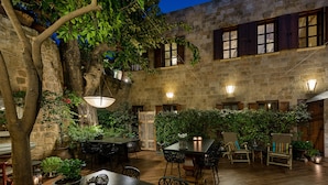 A courtyard with stone walls, wooden shutters, and hanging plants. There are tables and chairs arranged for outdoor dining. - Kókkini Porta Rossa (Rhodes)