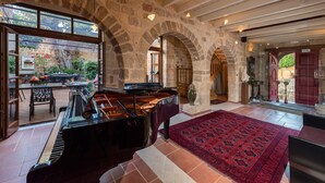 A grand piano in a spacious room with stone arches, a red patterned rug, and a view of an outdoor patio with tables and chairs. - Kókkini Porta Rossa (Rhodes)