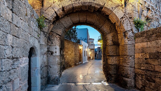 A narrow stone pathway leading through an arched stone gateway, flanked by buildings with traditional architecture. - Kókkini Porta Rossa (Rhodes)