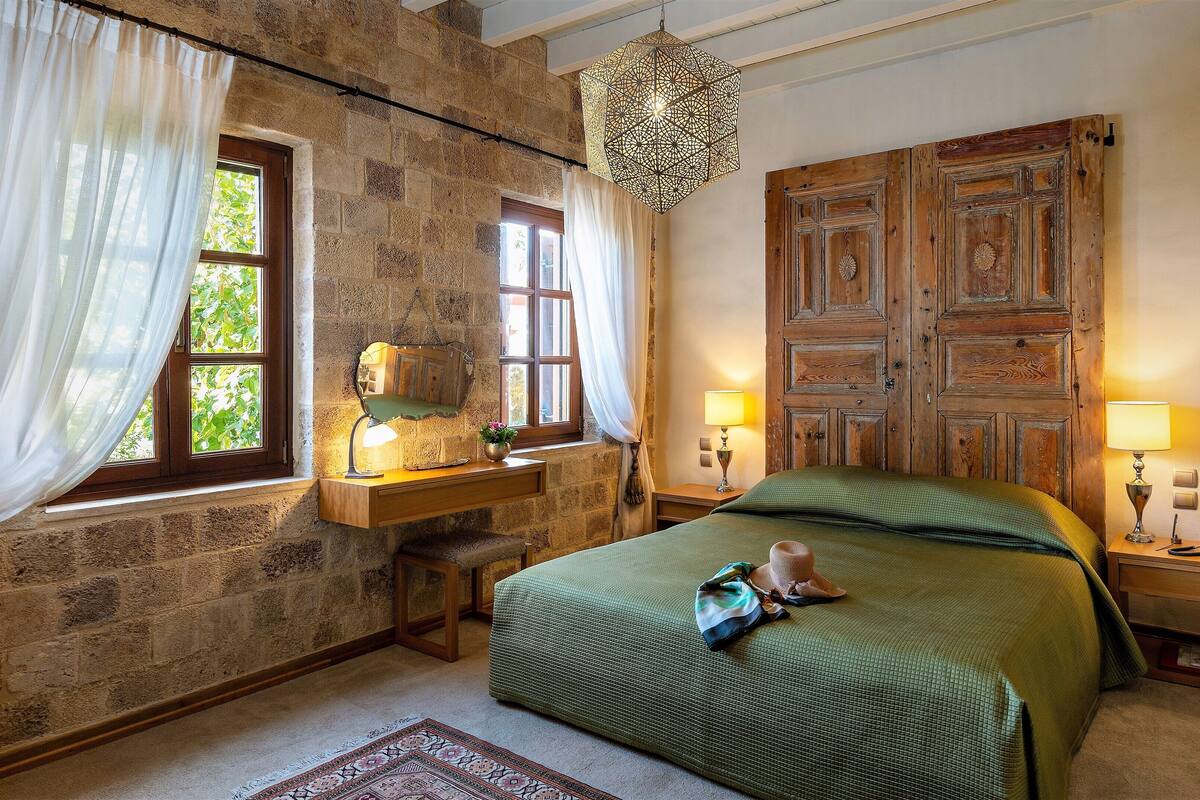 A bedroom with a stone wall, a wooden bed, two bedside tables with lamps, and a hanging light fixture.