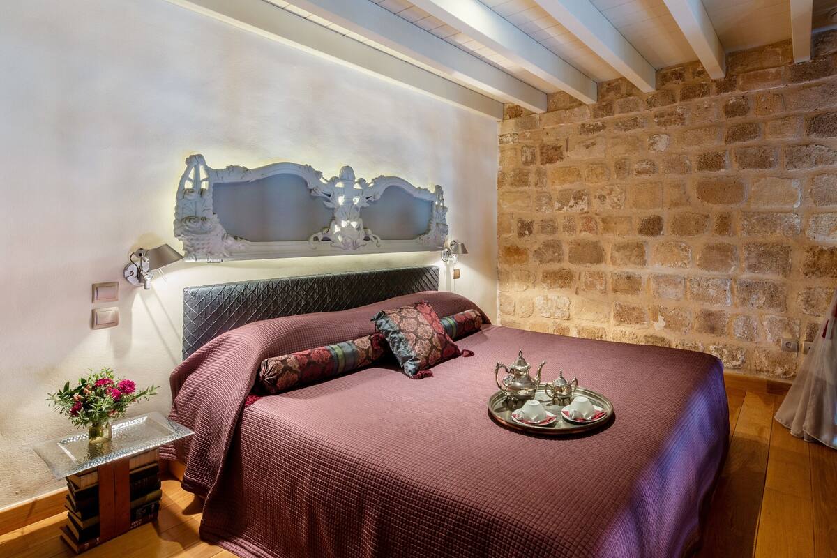 A bedroom with a stone wall, a bed with a headboard, a bedside table with a lamp, and a window with curtains.