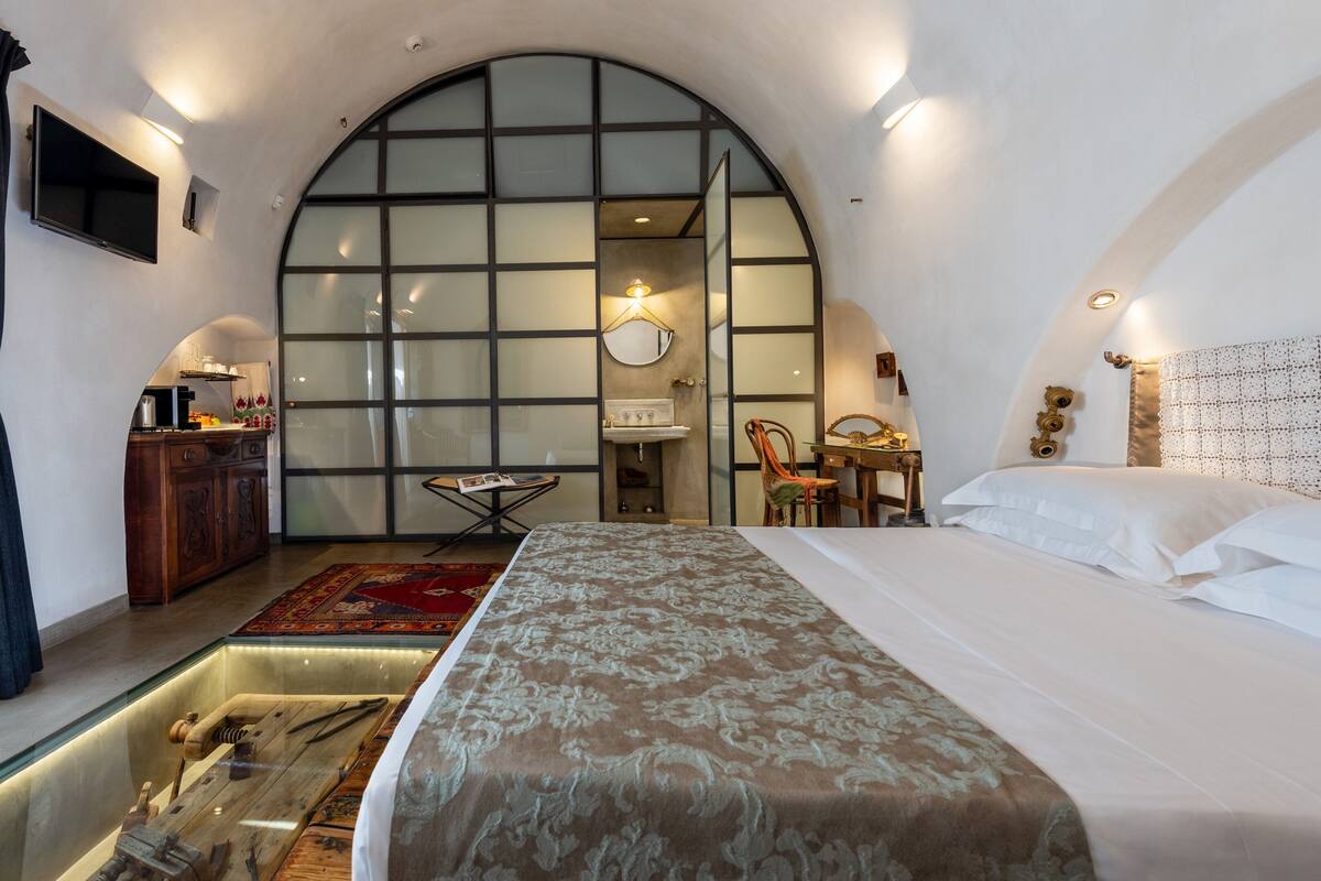 A modern hotel room with a large bed, a glass-walled bathroom, a desk, and a small table.