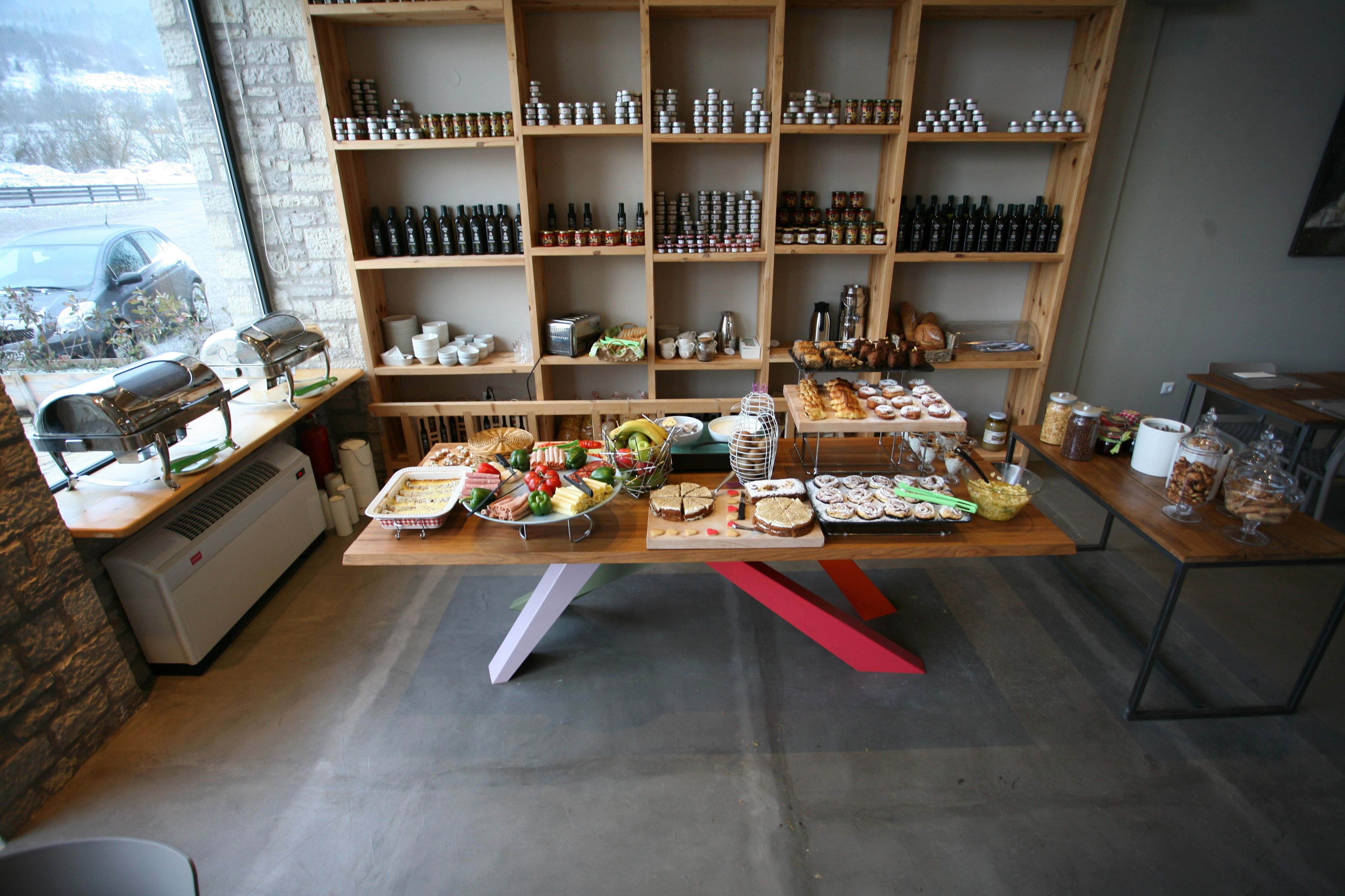 A bakery with a variety of baked goods displayed on tables, a large window with a view of the street, and shelves stocked with jars and bottles.