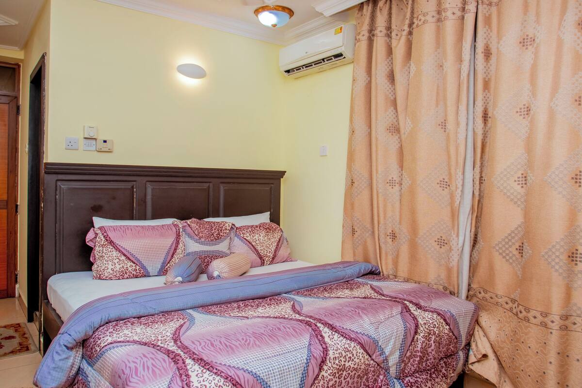 A neatly made bed with patterned bedding, a wooden headboard, and a wall-mounted air conditioner.