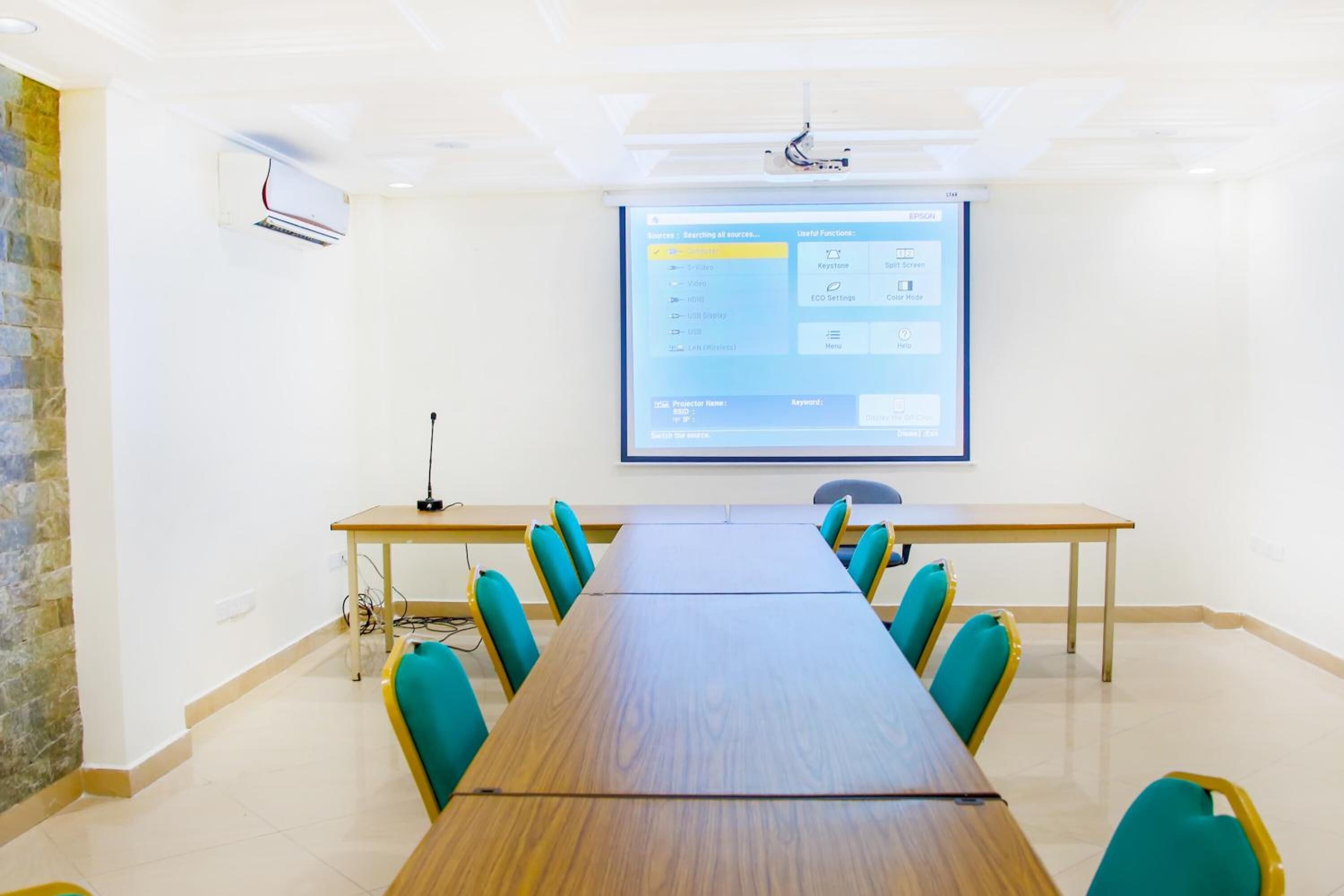 A conference room with a long table, teal chairs, a projector screen, and an air conditioning unit.