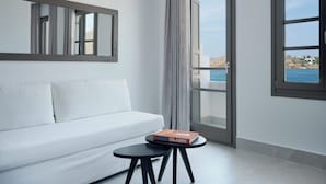 Une pièce moderne avec un canapé blanc, une petite table ronde sur laquelle repose un livre, et de grandes fenêtres offrant une vue sur la mer et les bâtiments.