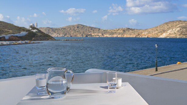 Une table est dressée sur une nappe blanche, près du bord de l’eau. On y trouve une carafe, deux verres et une tasse. En arrière-plan, on aperçoit des collines et un phare.