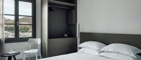 A modern hotel room with a bed, a small table with a cup and glass, a chair, and a wardrobe with a view of the city.