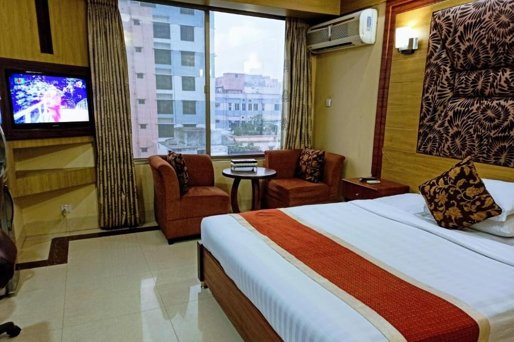 Habitación de hotel con cama, dos sillones, una mesita, televisor y vistas a la ciudad a través de amplios ventanales.