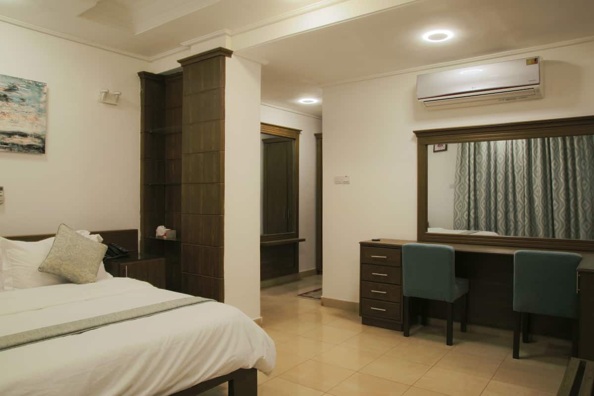 Habitación de hotel con cama, escritorio y dos sillas, aire acondicionado, armario y espejo con cortinas.