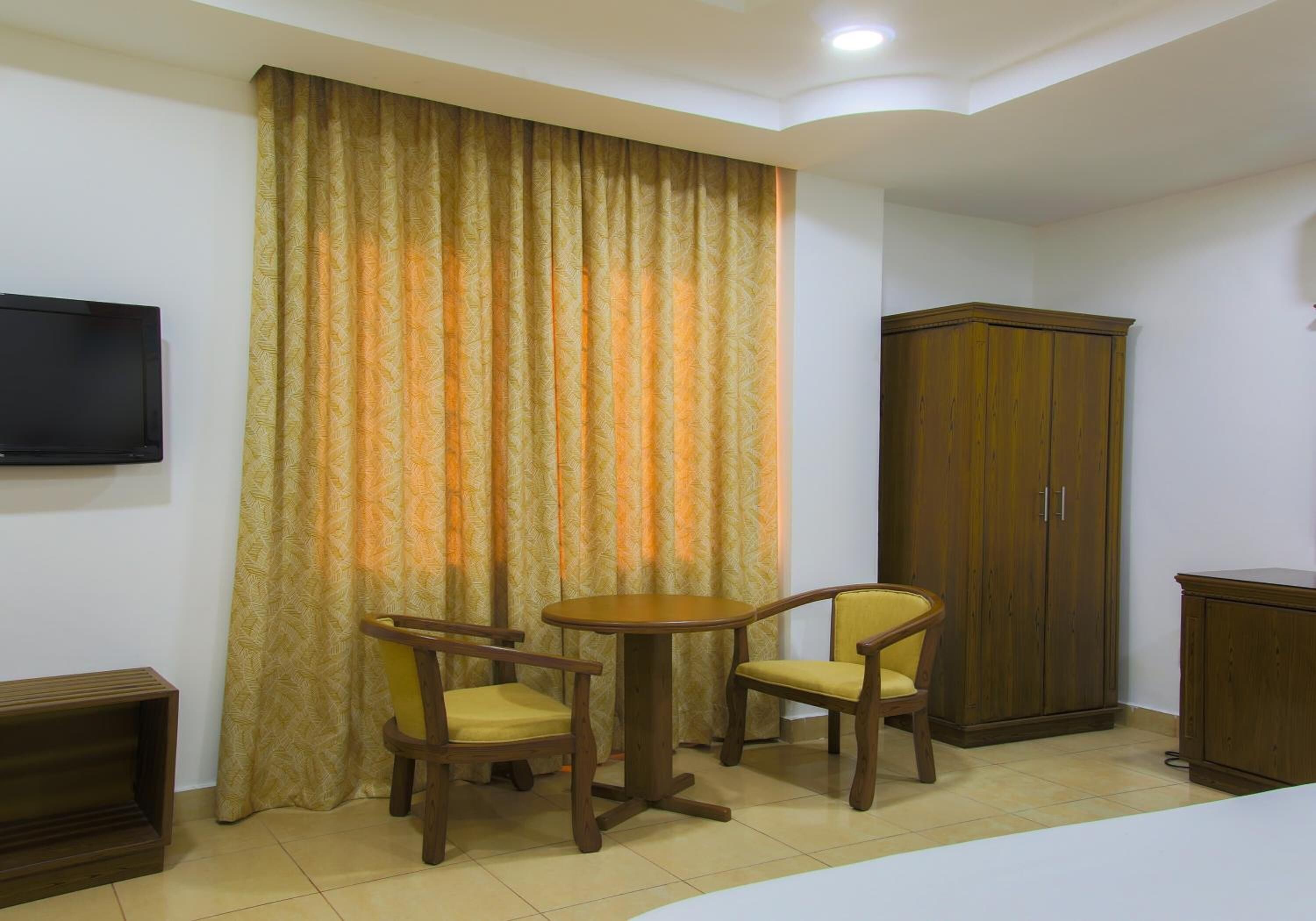 Una habitación con una televisión montada en la pared, un armario de madera, una mesa redonda con dos sillas y una cortina grande con diseño de hojas.