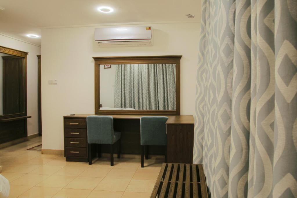 Una habitación con escritorio, dos sillas, un espejo grande, equipo de aire acondicionado y cortinas estampadas.