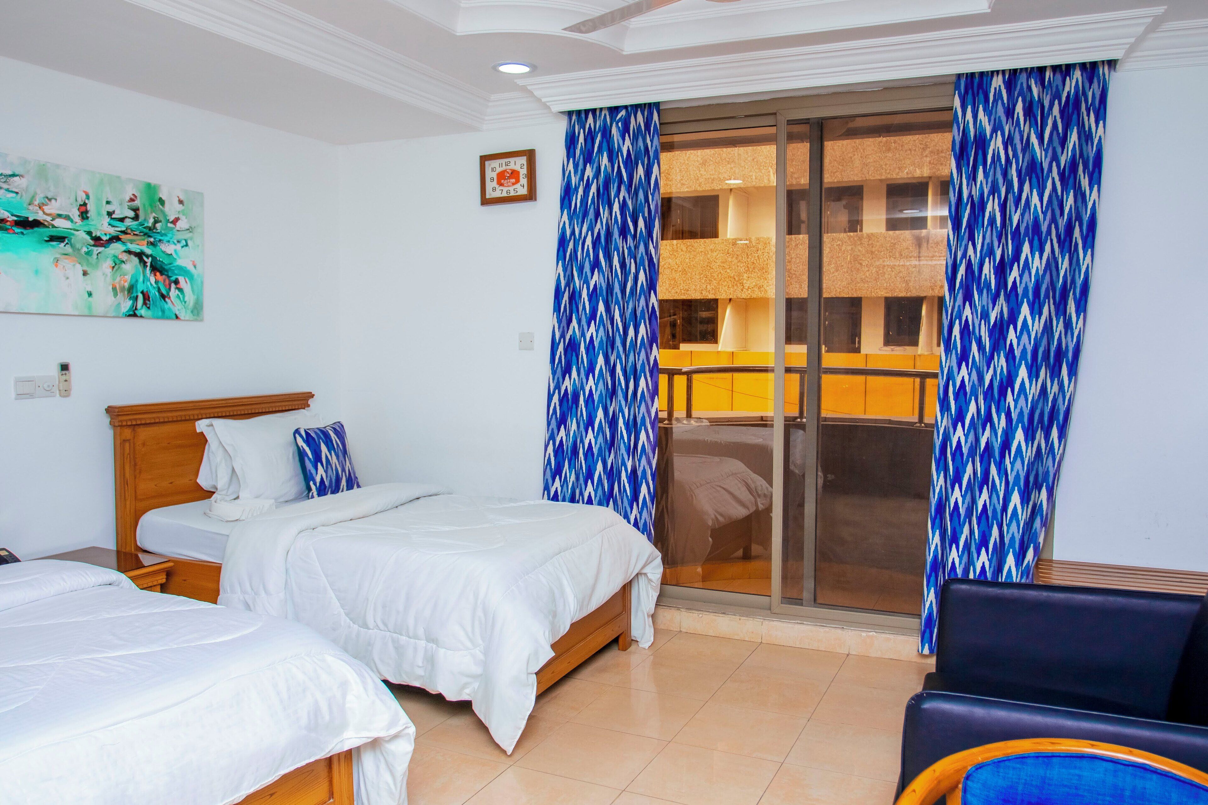 A hotel room with two beds, a large window with blue patterned curtains, a wooden headboard, and a painting on the wall.