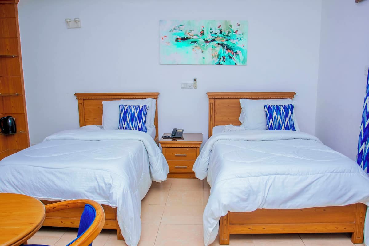 Habitación de hotel con dos camas, cabecera de madera, mesita de noche pequeña con teléfono, un cuadro en la pared y una silla de madera.