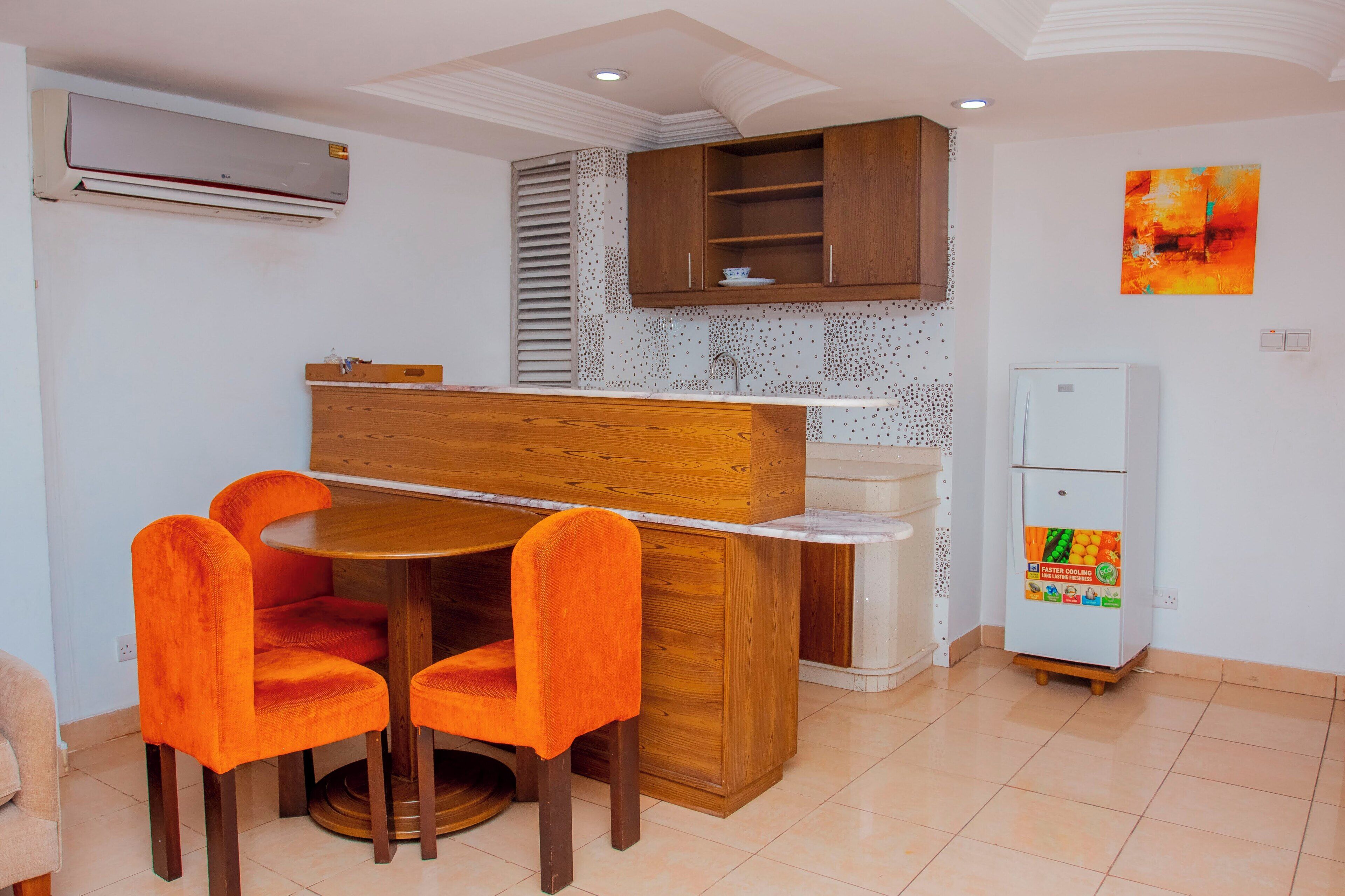 A compact kitchen with a wooden dining table and orange chairs, a white refrigerator, and a patterned backsplash.