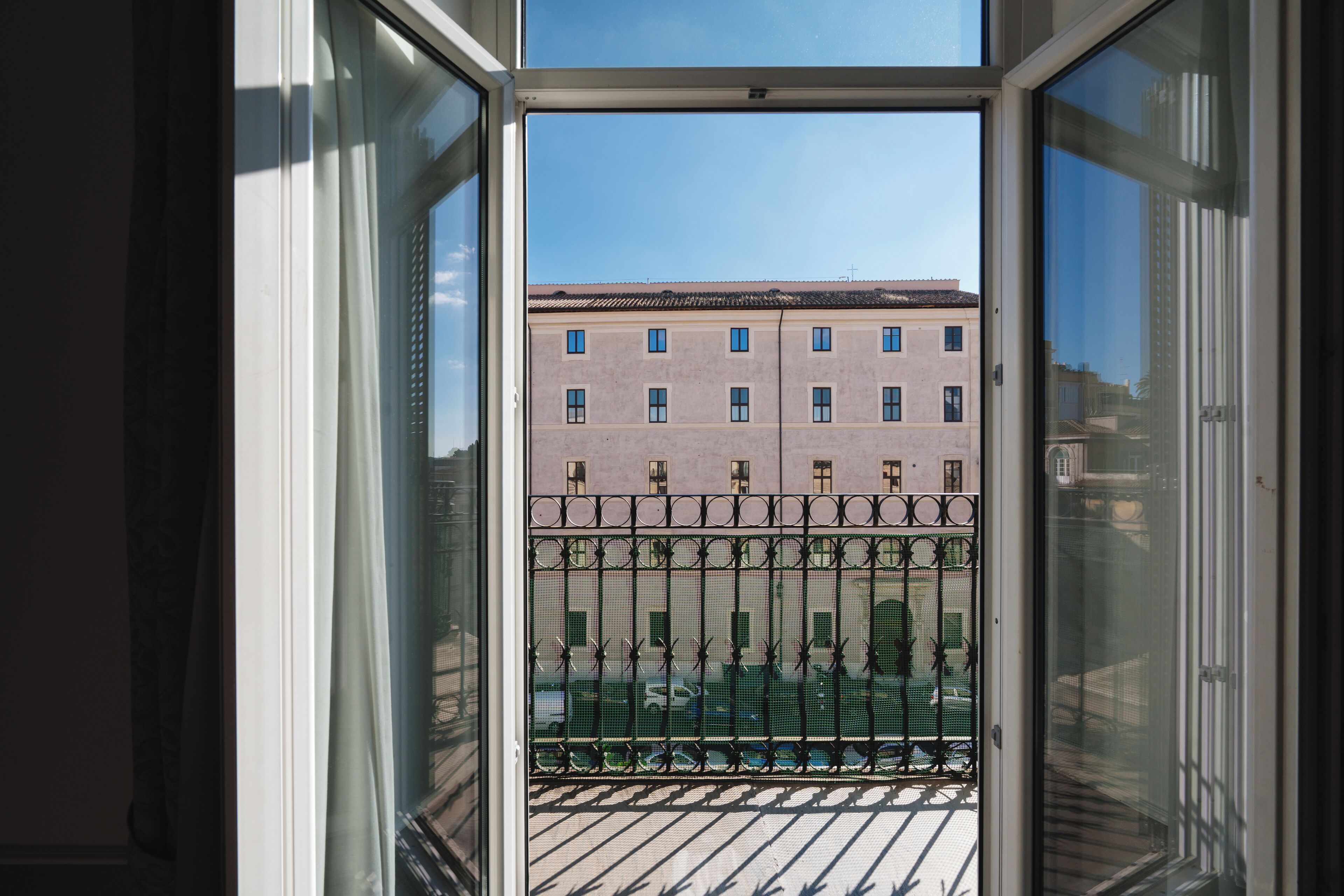 A view through a window with a metal balcony railing, a building with multiple windows, and a clear sky.