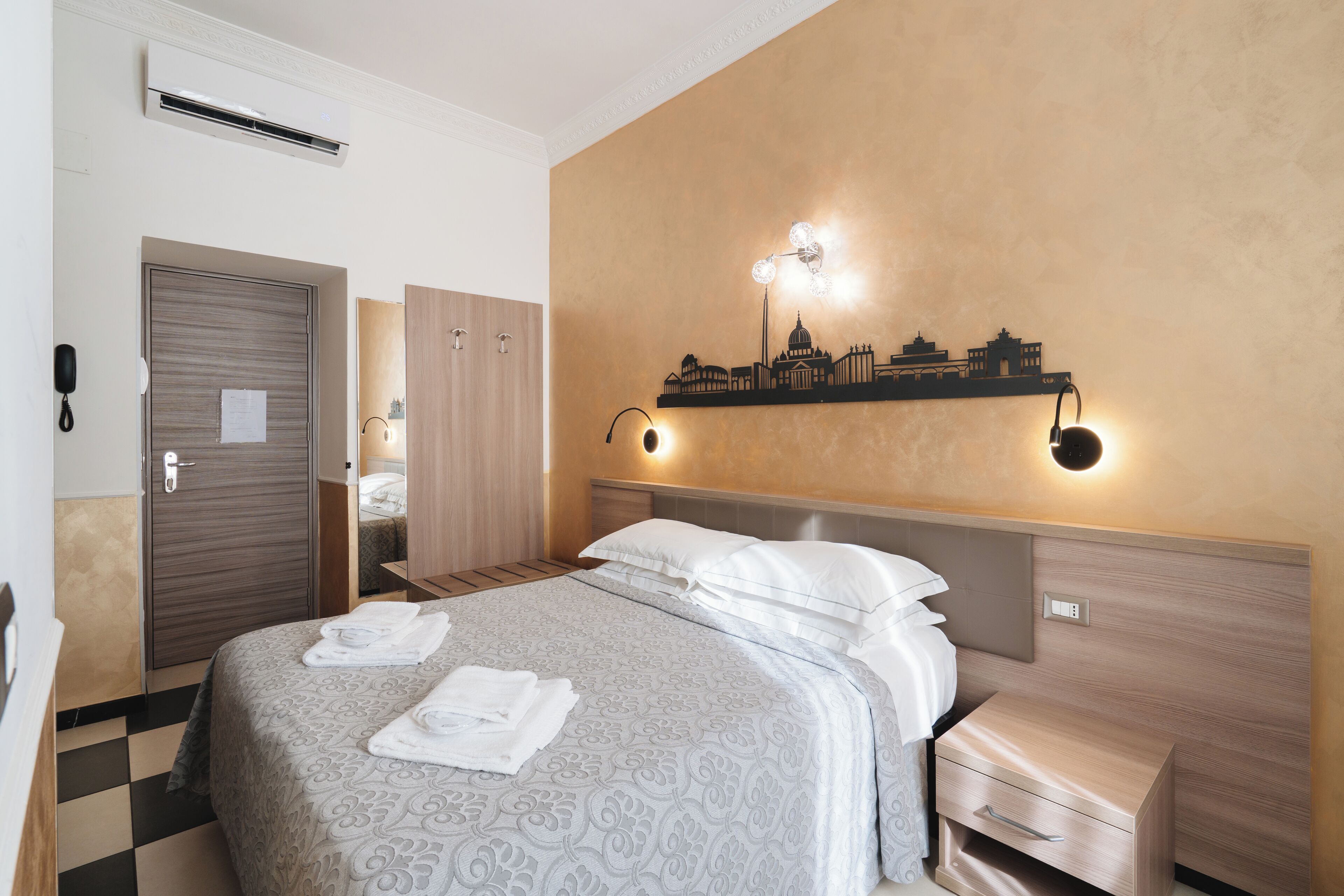 A hotel room with a bed, bedside tables, a wall-mounted shelf with a cityscape decoration, and a door with a phone and a note.