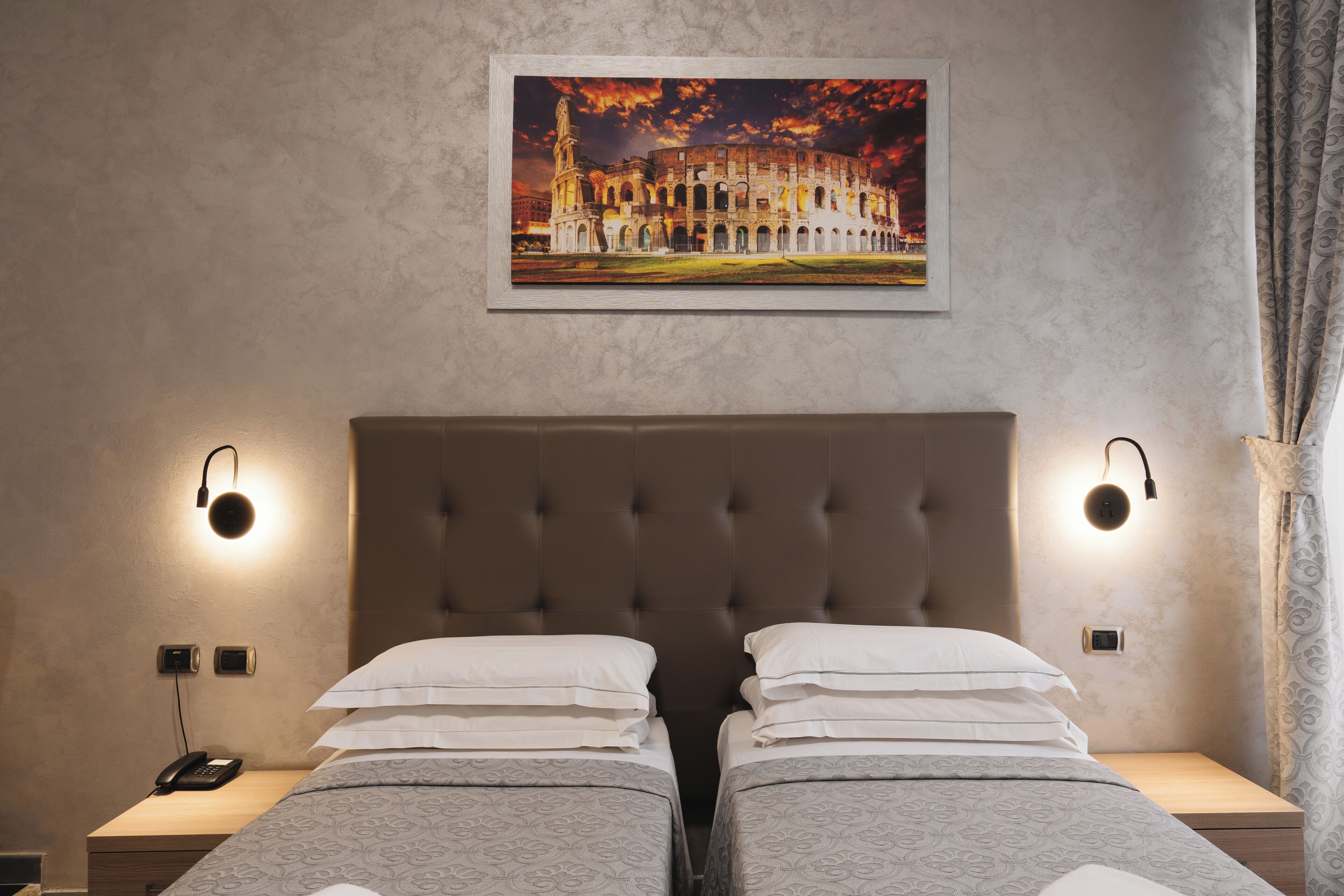 Habitació d'hotel amb un llit gran, dues taules de nit, una petita taula, un telèfon i un quadre amb una imatge d'un edifici històric a la paret.