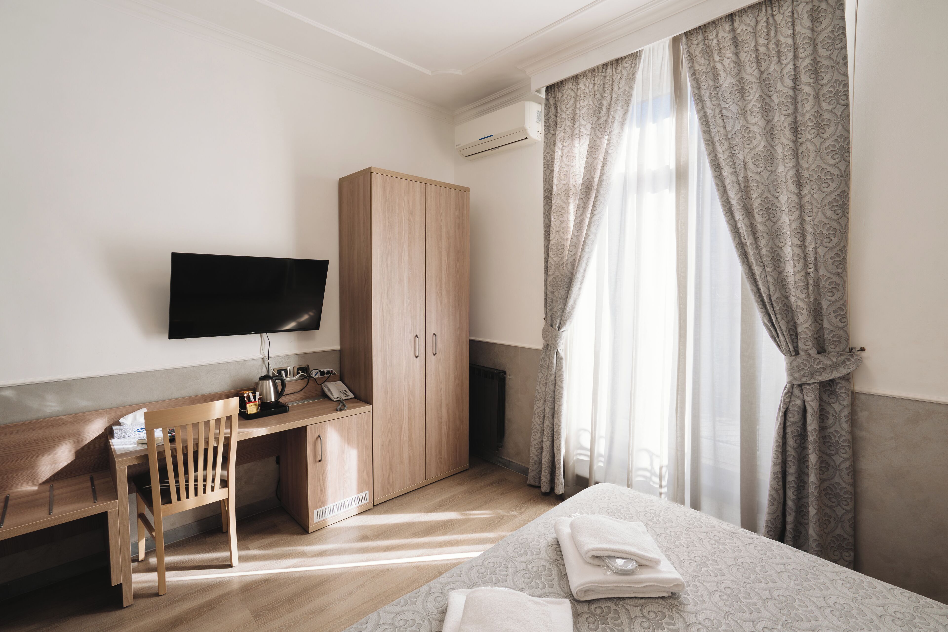 A hotel room with a wooden desk, a flat-screen TV, a wardrobe, a bed with neatly folded towels, and large patterned curtains.