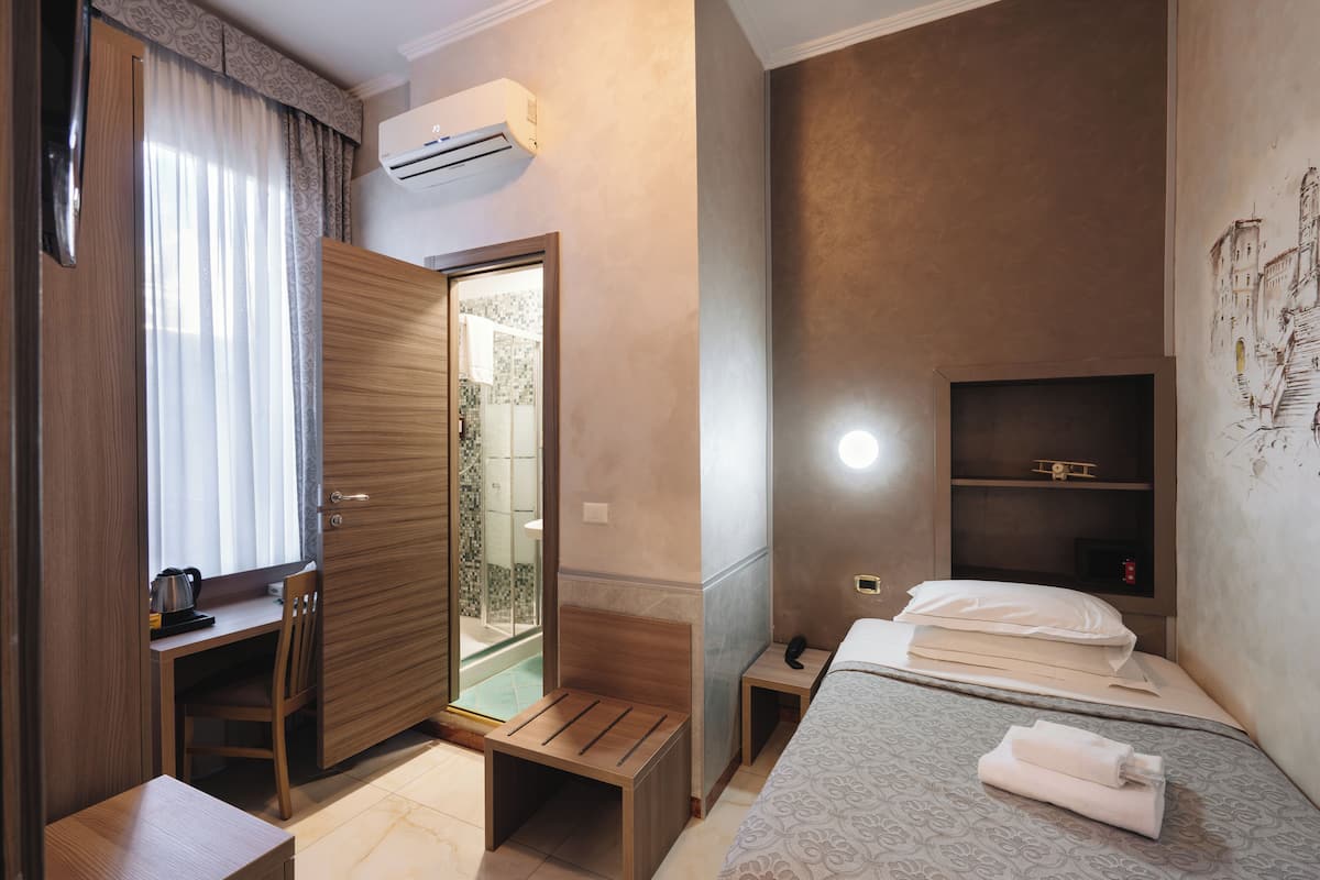 Habitación de hotel con un escritorio de madera, una cama con toallas ordenadas, una mesita de noche con un teléfono y un estante en la pared.