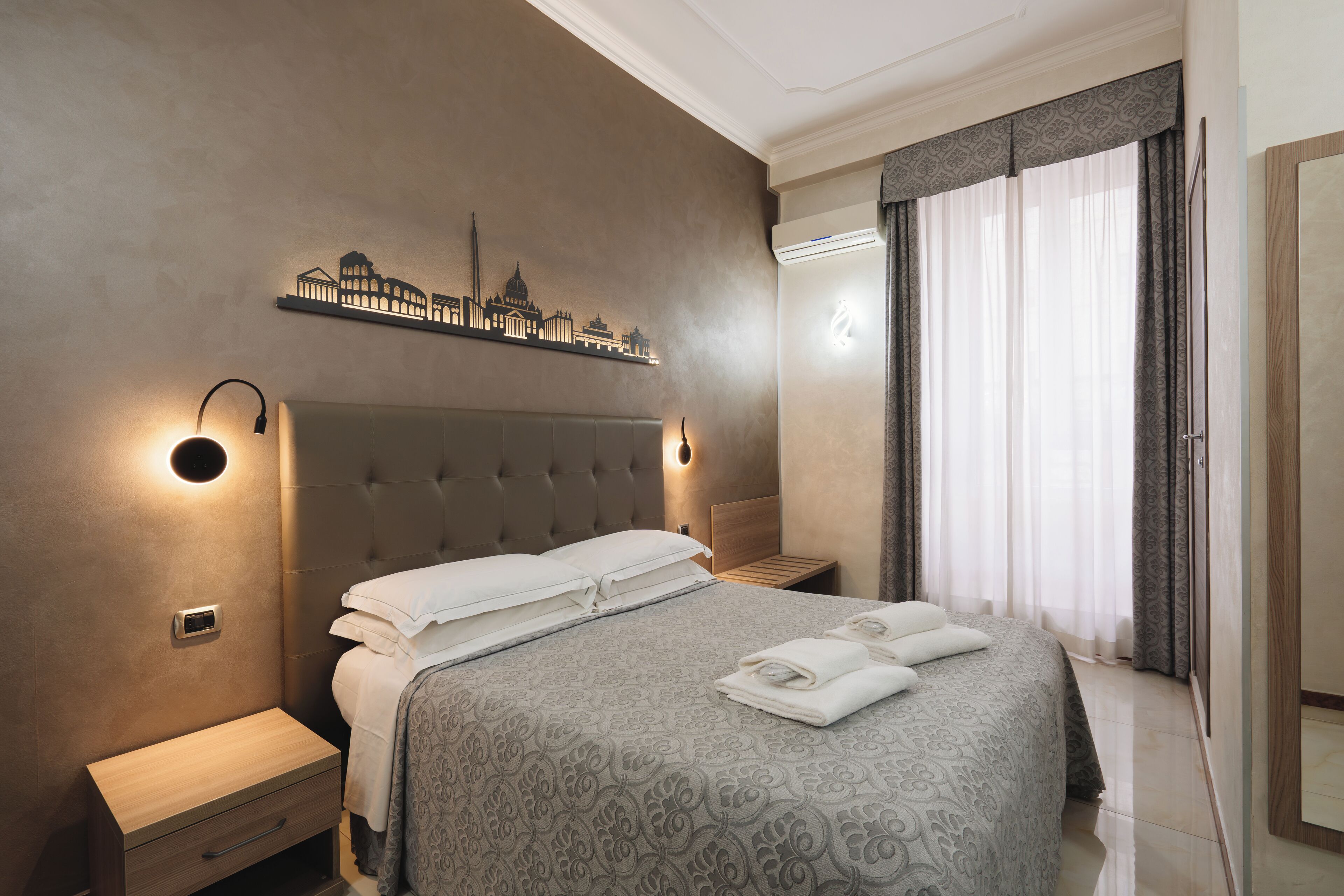Habitació d'hotel moderna amb un llit ben fet, un quadre mural amb un paisatge urbà, una taula de nit i un gran finestral amb cortines.