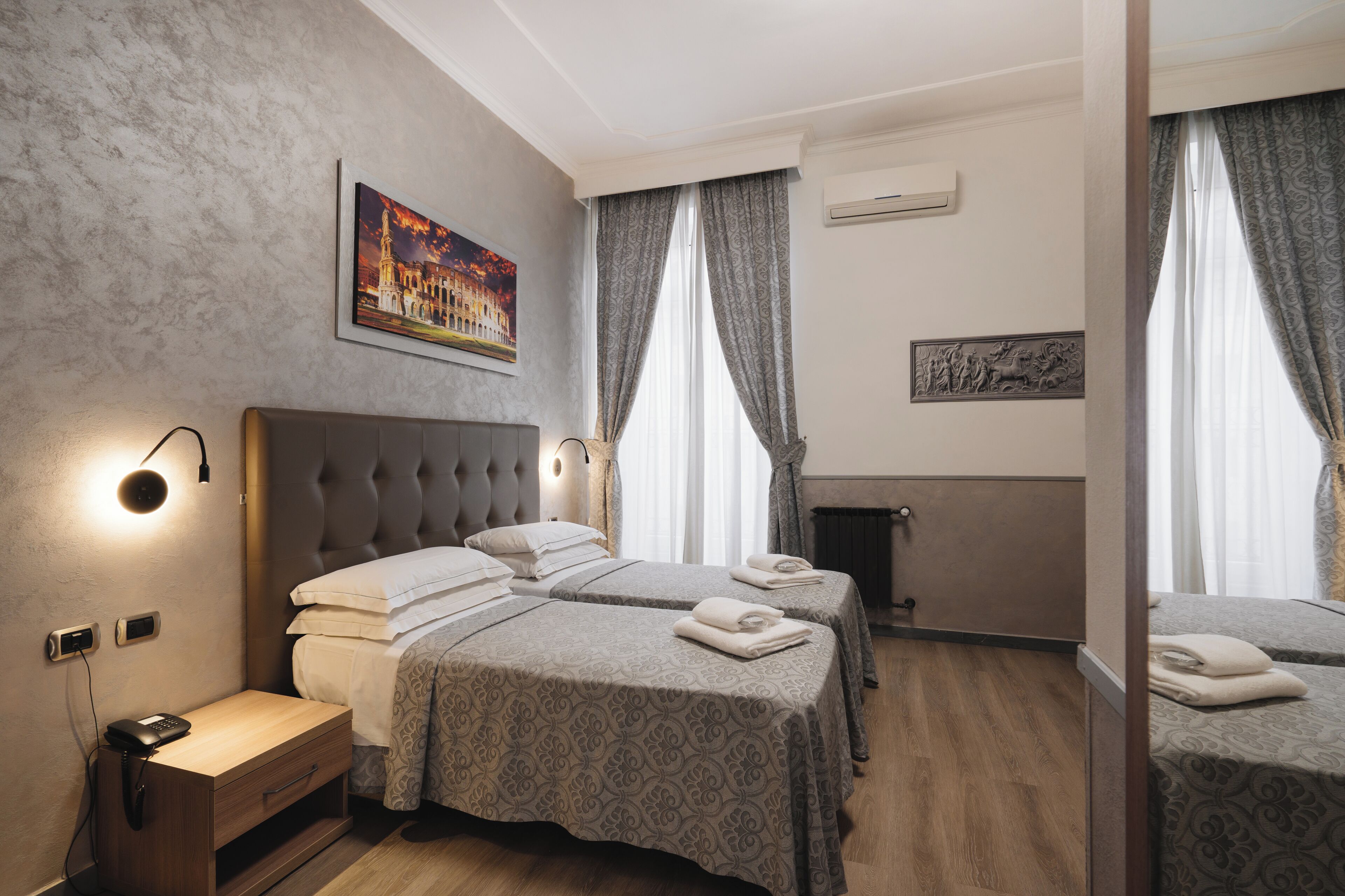 Camera d'albergo con due letti, un comodino, un condizionatore d'aria a muro e un quadro raffigurante un edificio.
