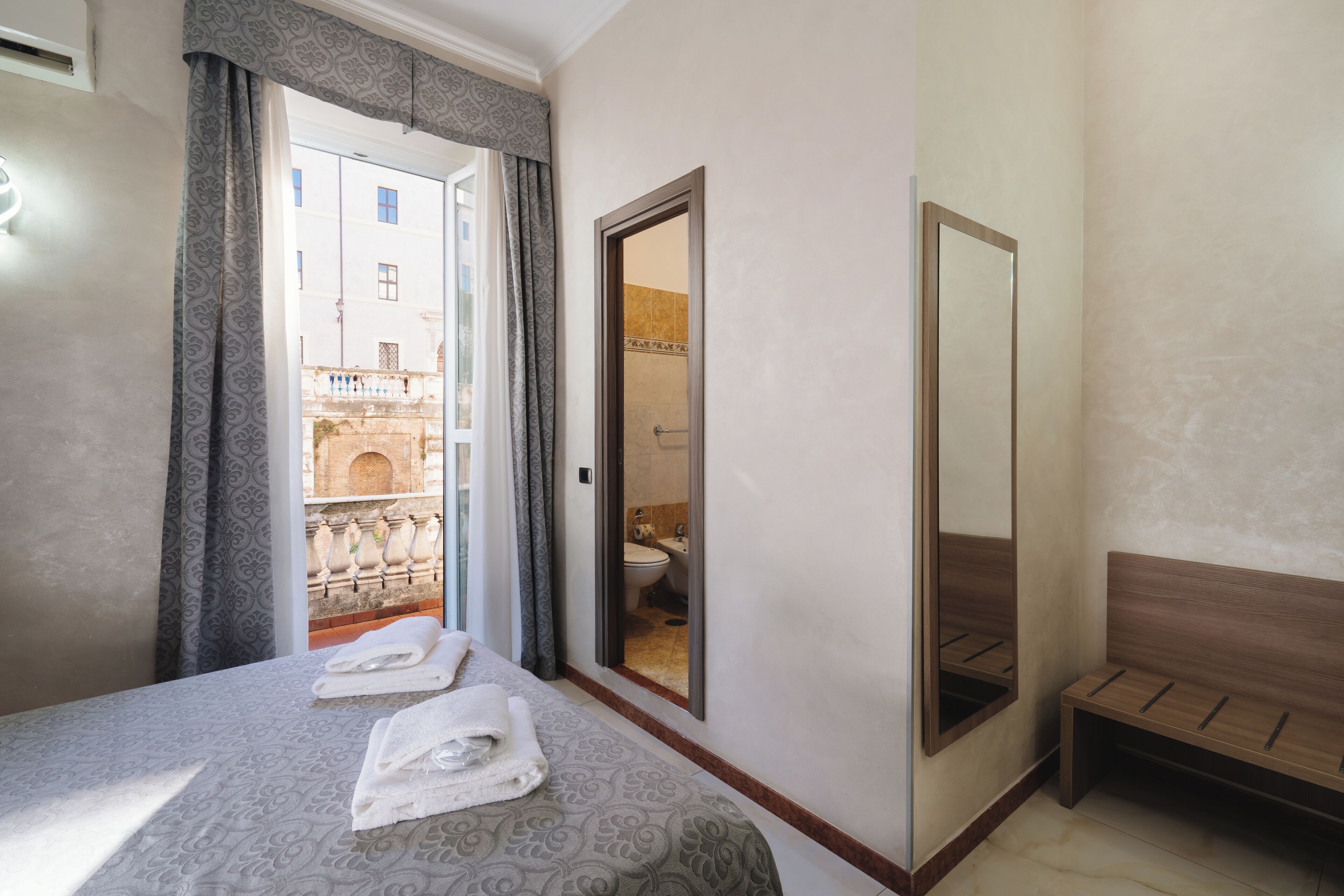 A hotel room with a bed, a bench, a mirror, and a view of the outside.