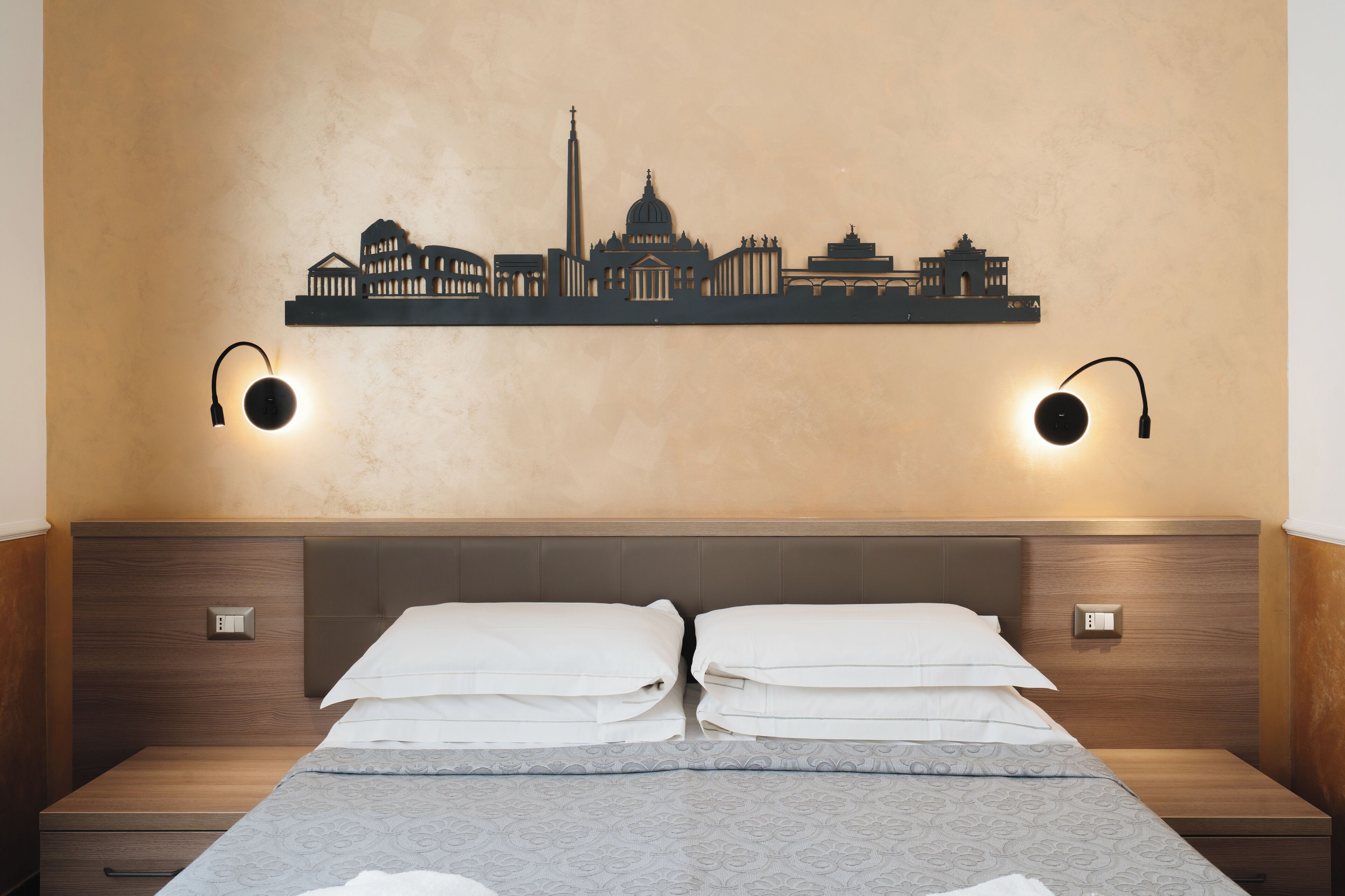 Une chambre d’hôtel comprenant un lit, deux tables de chevet, une œuvre d’art représentant un paysage urbain accrochée au mur, et deux lampes murales.
