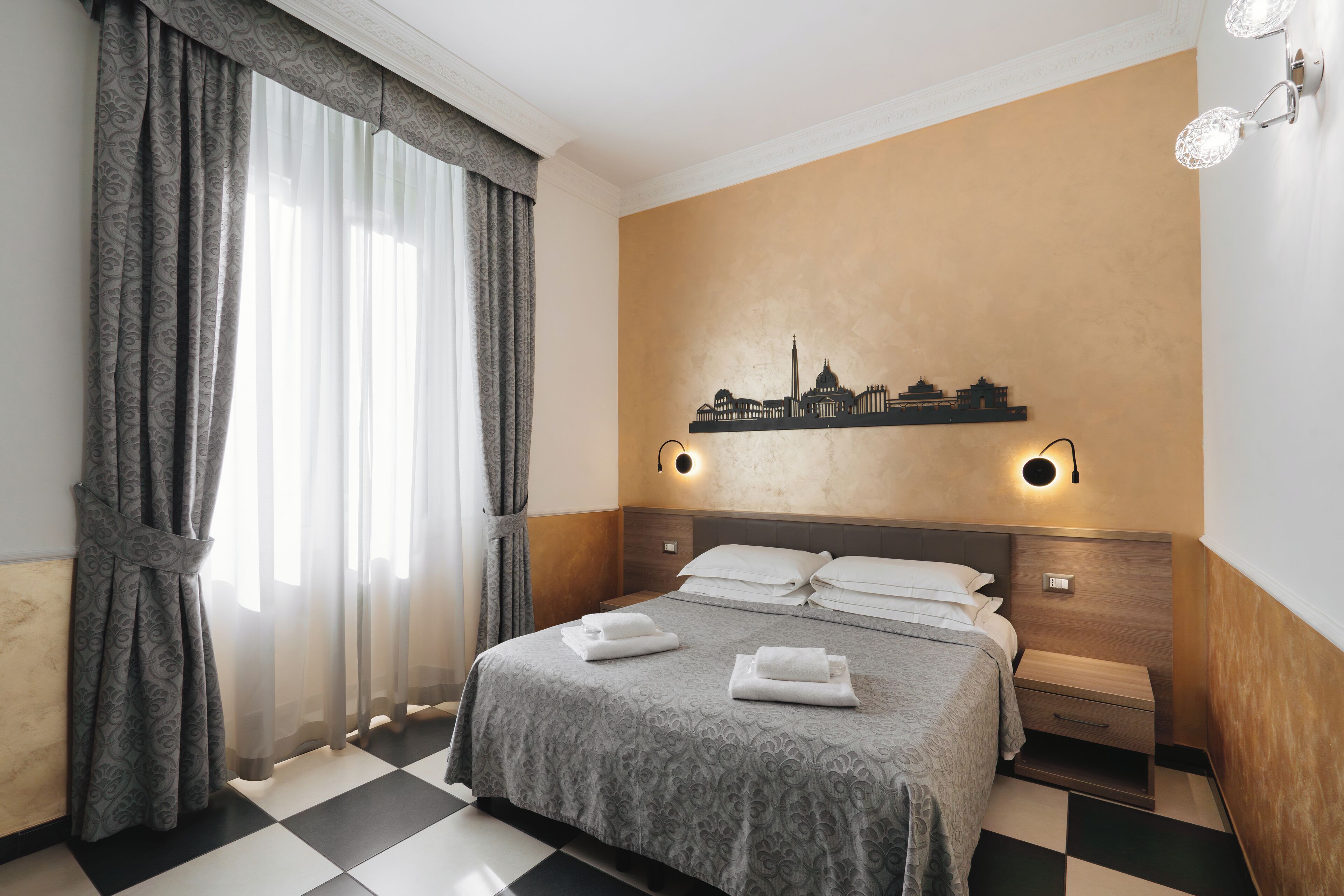 Habitación de hotel con una cama, una mesita de noche, un estante en la pared con un adorno de paisaje urbano y una ventana con cortinas translúcidas.