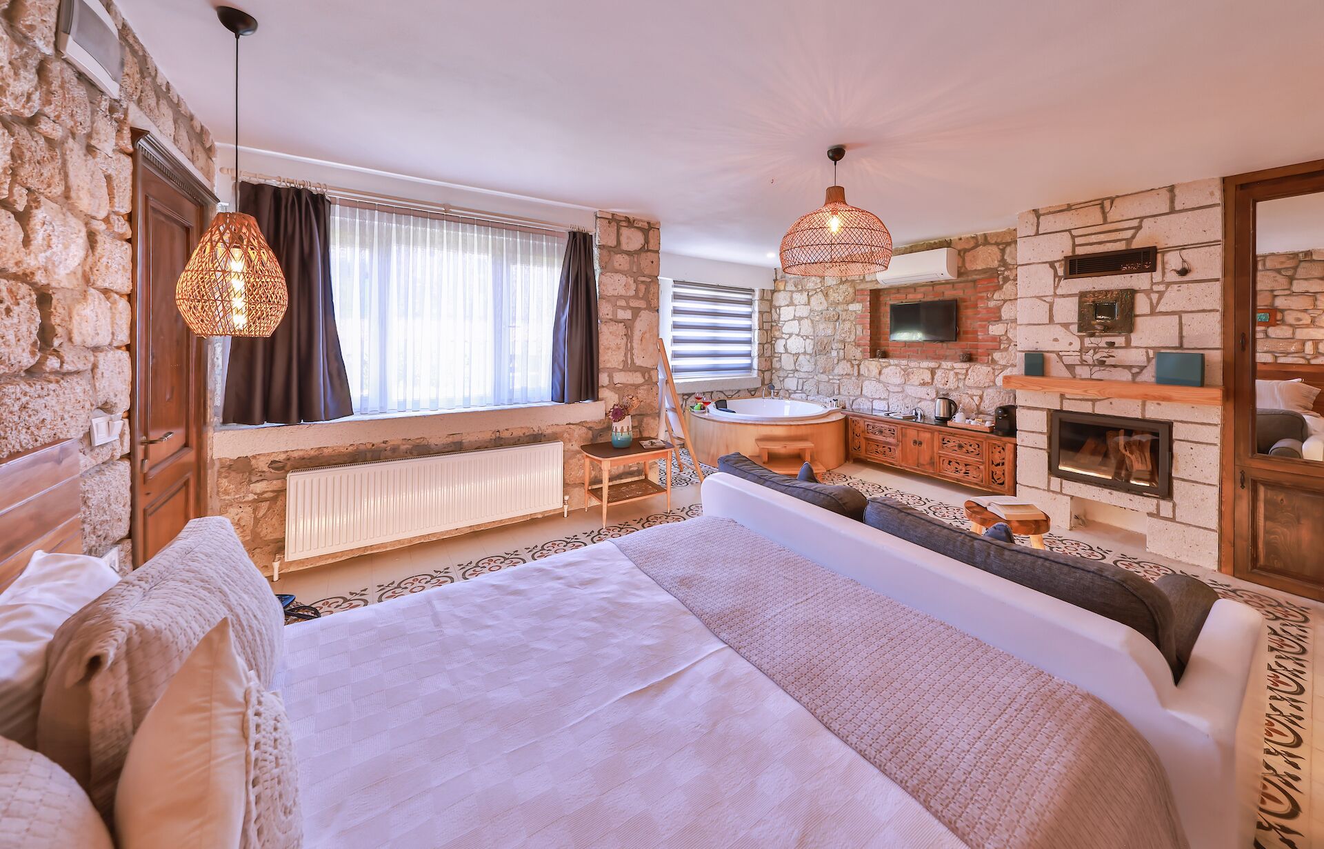 A bedroom with a large bed, a sitting area with a sofa and a small table, a stone wall, a fireplace, and a window with curtains.