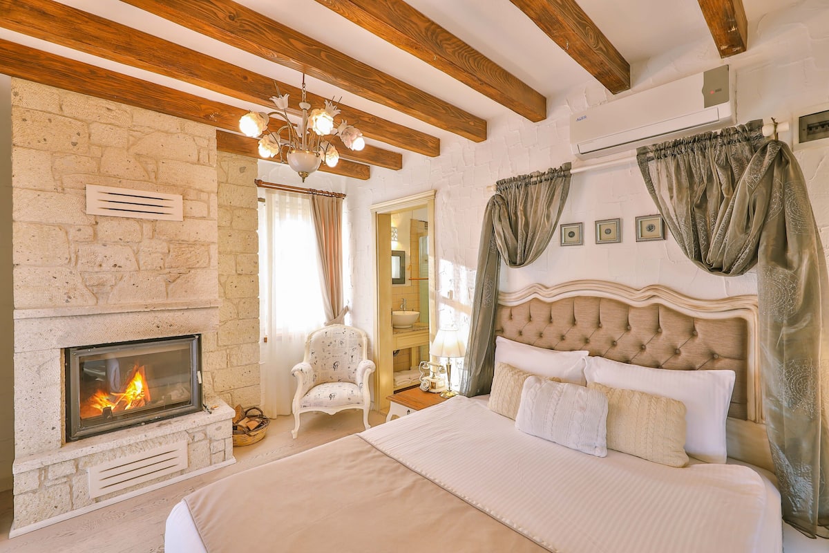 A bedroom with a fireplace, a bed with multiple pillows, a chair, and a nightstand with a lamp.