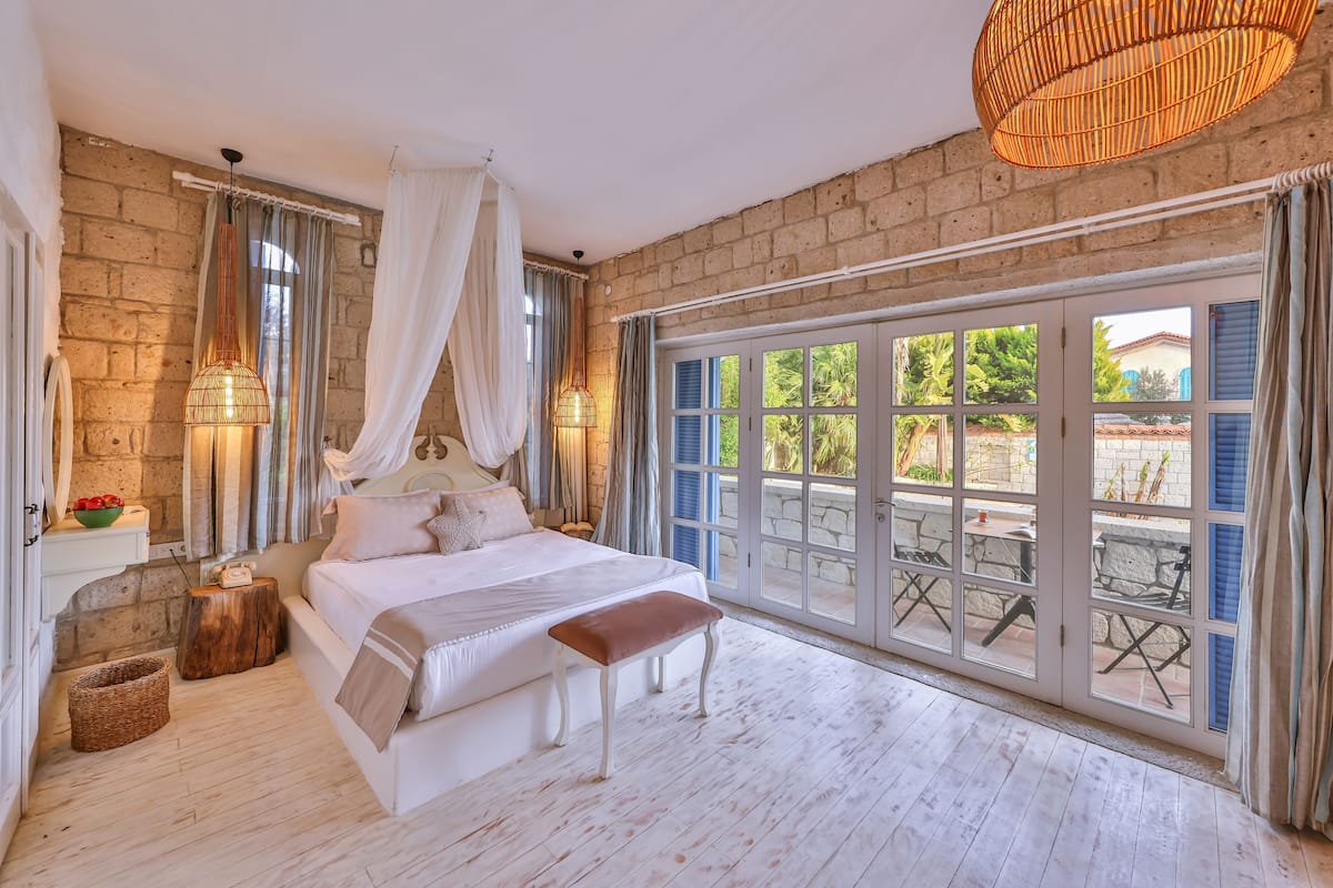 A bedroom with a large bed, a bench, a wicker hanging light, and a view of the outside through large glass doors.