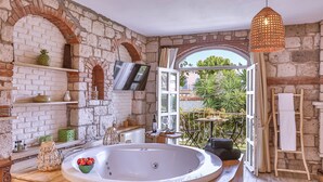 Un baño con una bañera independiente, escalones de madera, paredes de piedra y vistas a la vegetación exterior.