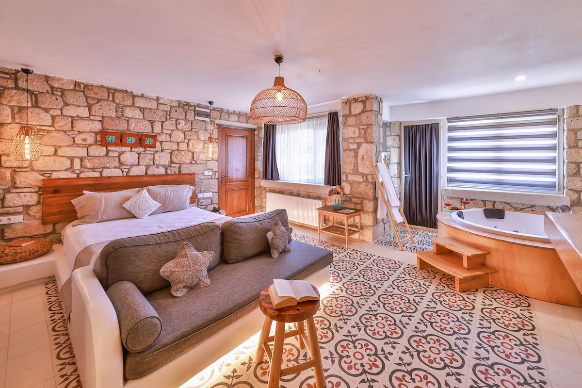 A cozy bedroom with a stone wall, a wooden bed, a grey sofa, a small table, and a bathtub.