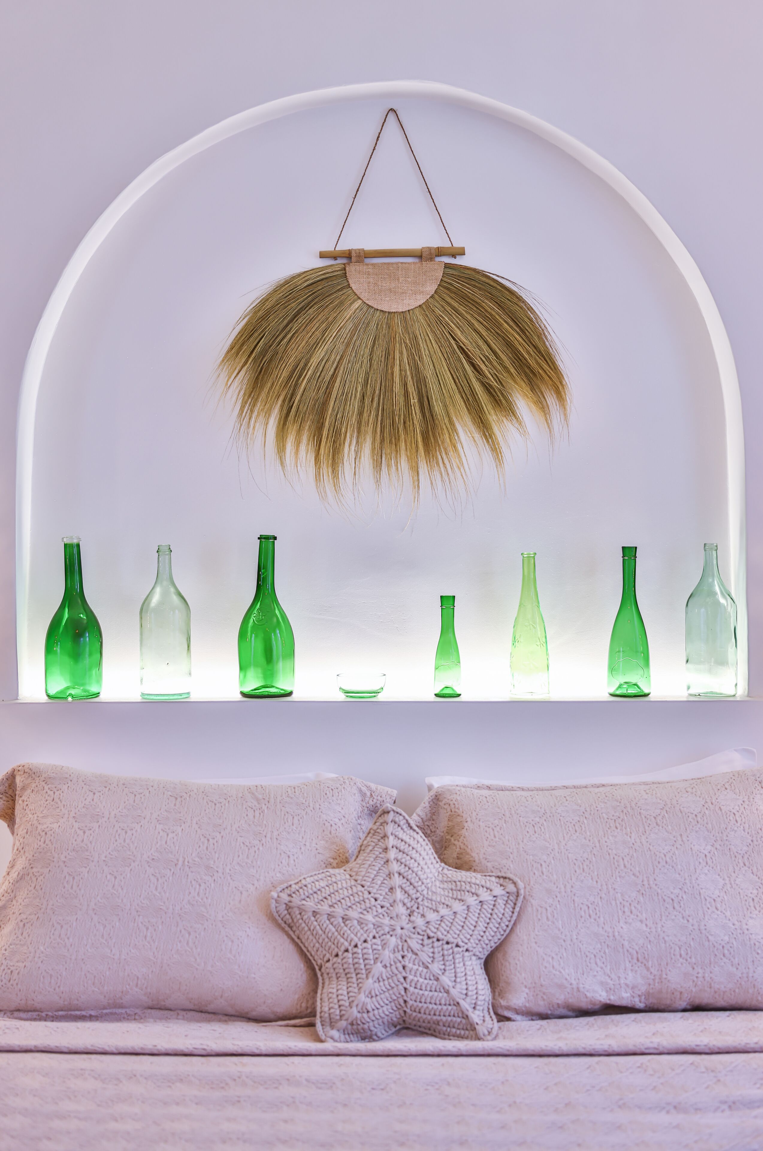 Una habitación con un ventanal arqueado blanco que muestra botellas verdes, un tapiz tejido y un cojín con forma de estrella sobre un sofá color beige.