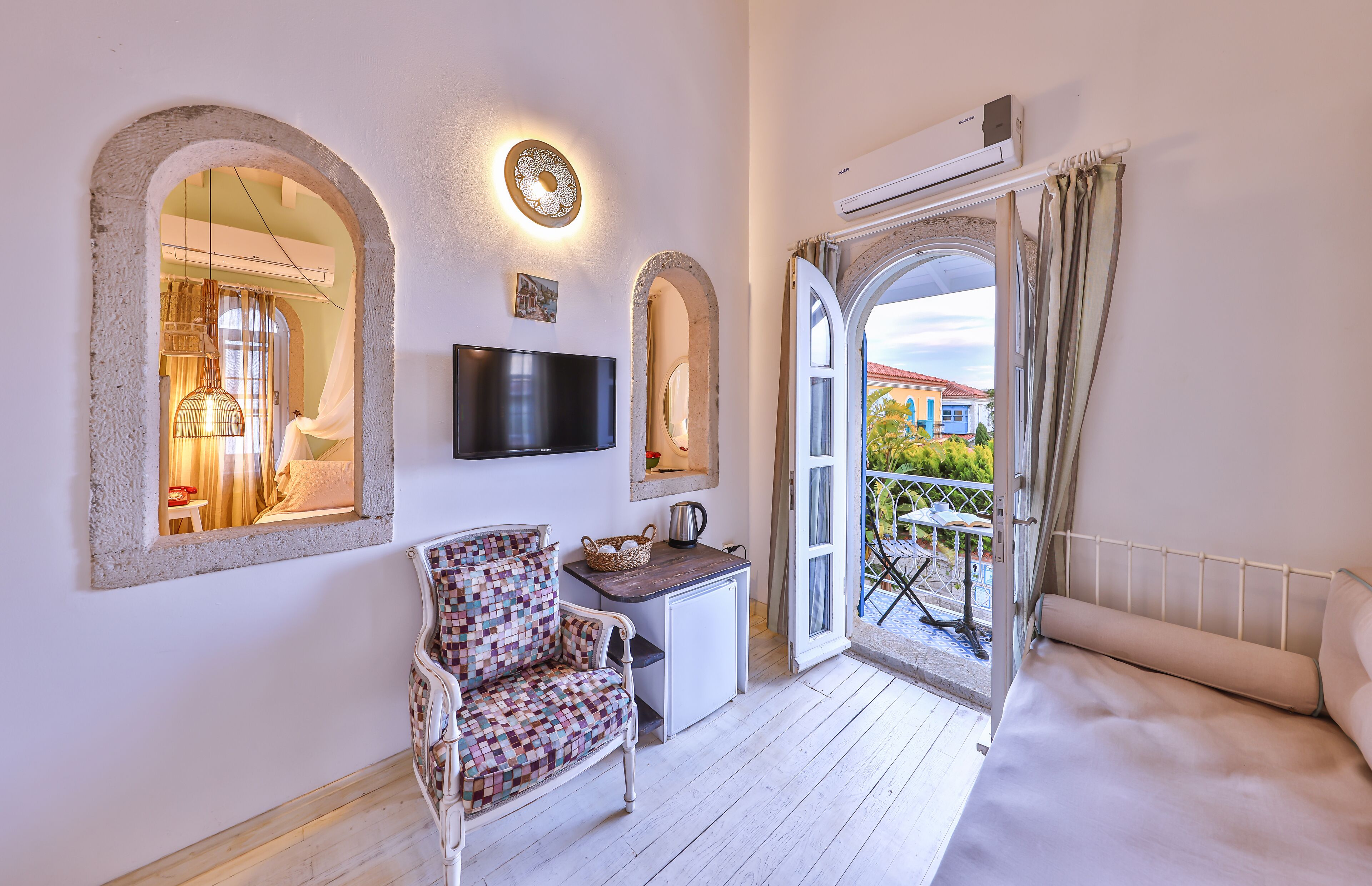 Habitación con sofá, un sillón estampado, una mesita con hervidor, televisión de pantalla plana y balcón con vistas a edificios.