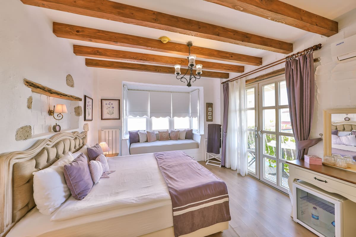 A bedroom with a large bed, wooden beams, a chandelier, a window with curtains, and a bedside table with a lamp.