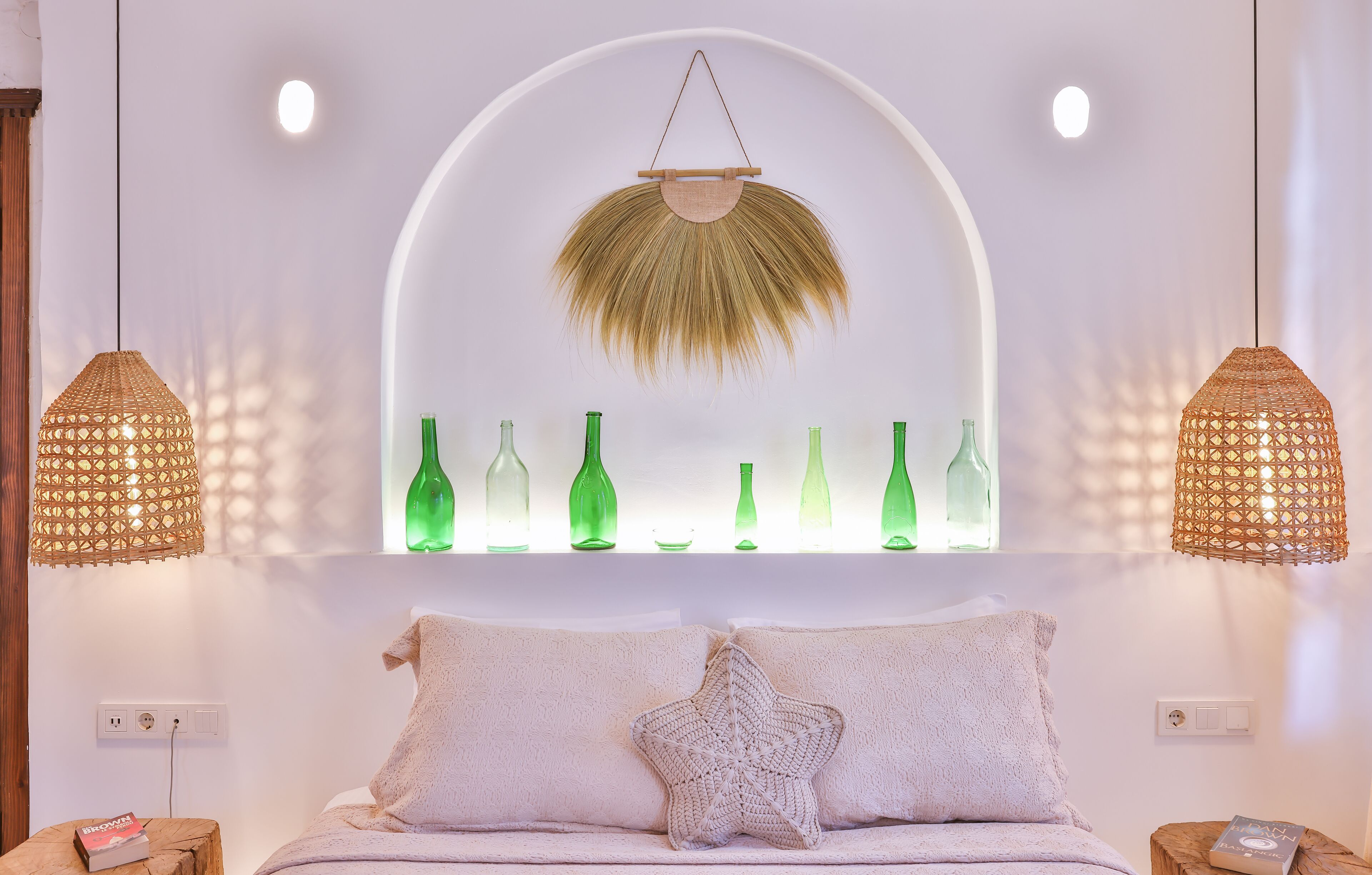 Een moderne slaapkamer met een witte muur, een groot raam, groene flessen, een witte bank met een stervormig kussen en twee hanglampen.