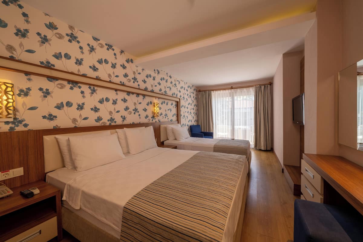 A hotel room with two beds, a wooden nightstand with a phone and remote, a desk with drawers, a television, and a window with curtains.