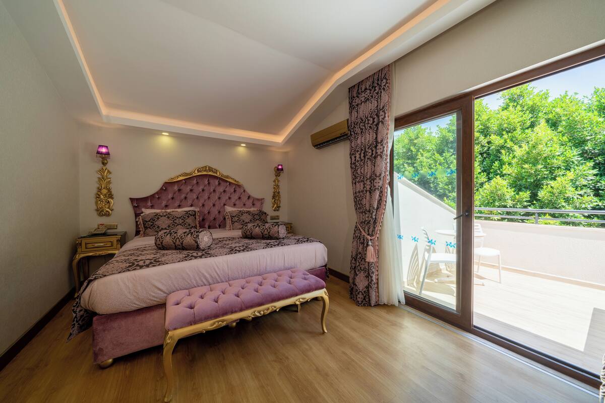 A bedroom with a large bed, a bench, a nightstand, and a window with a view of greenery.