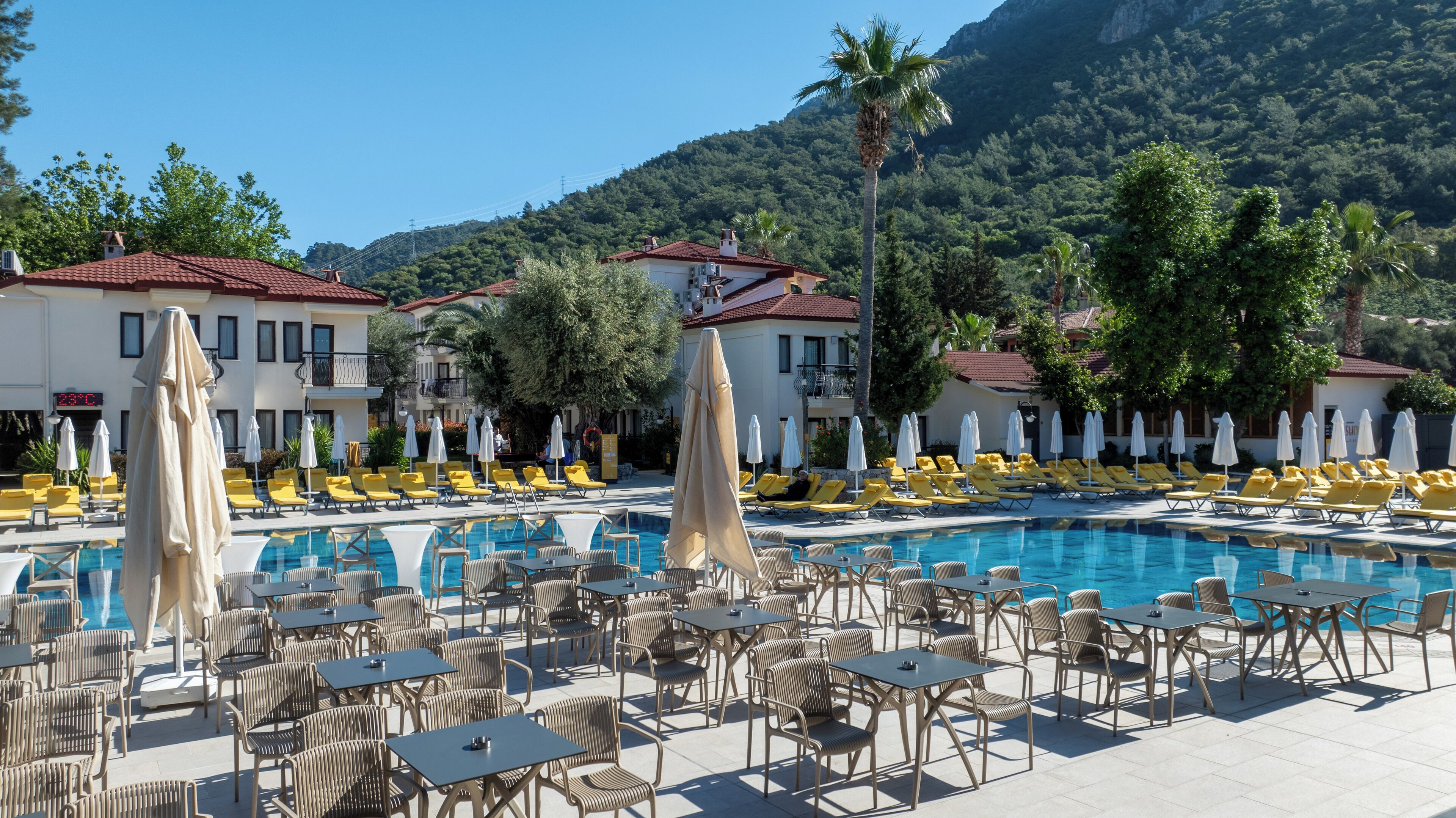 A poolside area with lounge chairs and umbrellas, a pool, and buildings with red roofs.